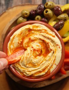 overhead shot of dunking red pepper into bowl of hummus with peppers, celery and olives blurred in background