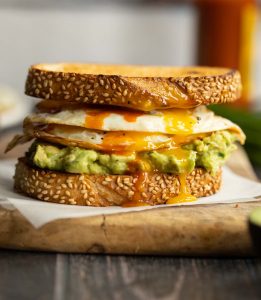 sandwich on parchment paper on chopping board with chives, hot sauce and eggs blurred in background