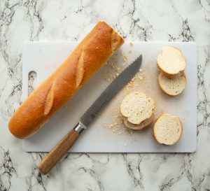 overhead shot of french bread being sliced on plastic chopping board with bread knife