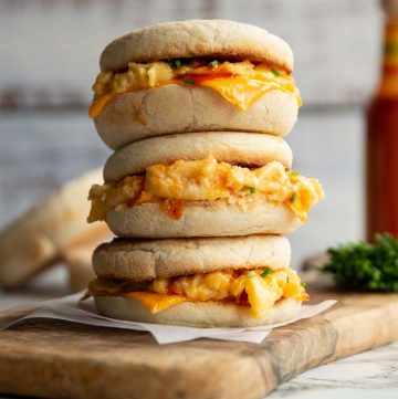 3 sandwiches stacked on each other on wooden board with muffins, chives and hot sauce blurred in background