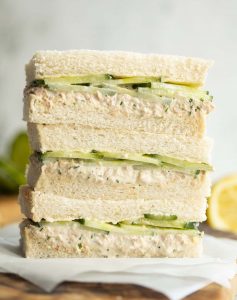 3 finger sandwiches stacked on each other on wooden board with lemon and cucumber blurred in background