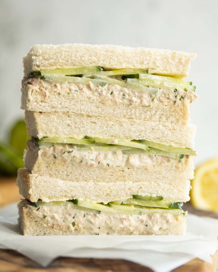 3 finger sandwiches stacked on each other on wooden board with lemon and cucumber blurred in background