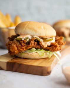 chicken sandwich on wooden board with sandwich and fries blurred in background and dip blurred at front