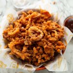 fried onions in wooden bowl with salt and bbq sauce blurred in background