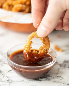 close up shot of hand dunking in crispy onion to small glass pot of bbq sauce