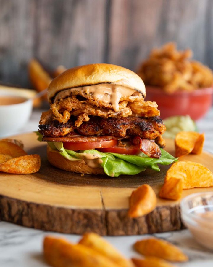 chicken sandwich on wooden board surrounded by potato wedges and dip