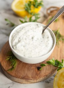 small white pot of tartar sauce on small white board surround by lemon, parsley and dill
