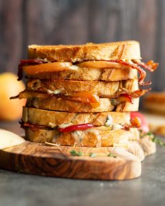 3 sandwiches stacked on each other on wooden board with apple and bread blurred in background