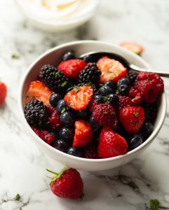 berries in small white bowl with silver spoon surrounded by strawberries on marble background