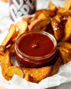 small glass pot of bbq sauce surrounded by potato wedges on parchment paper