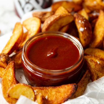 small glass pot of bbq sauce surrounded by potato wedges on parchment paper