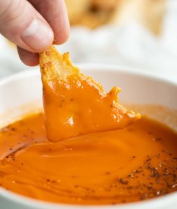 fingers dunking chip into small white bowl of tomato soup