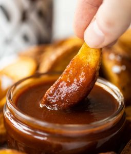 close up shot of fingers dunking potato wedges into glass pot of bbq sauce