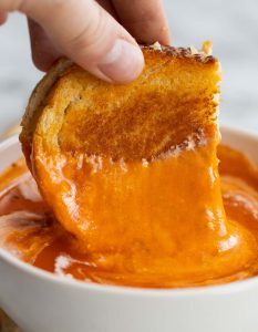 dunking grilled cheese into small white bowl of tomato soup