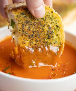hand dunking grilled cheese into bowl of tomato soup