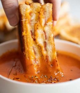 close up shot of hand dunking grilled cheese halfinto small white bowl of tomato soup