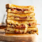 4 bacon grilled cheese halves stacked on each other on wooden board with maple syrup poured over