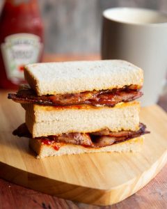 2 bacon sandwich halves stacked on each other with ketchup and cup of tea blurred in background