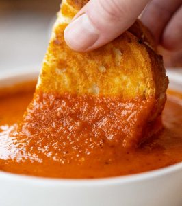 close up shot of hand dunking grilled cheese into small white bowl of soup