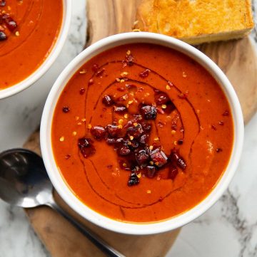 overhead shot of red pepper soup served in white bowl on wooden board with spoon and grilled cheese