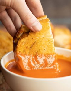 close up shot of hand dunking grilled cheese into small white bowl of tomato soup