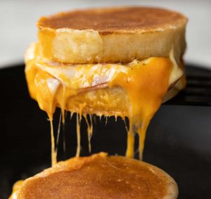 close up shot of black spatula lifting crumpet sandwich from pan with cheese dripping out