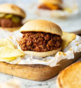 sloppy joe on wrapper on chopping board with crisps and two more blurred in background