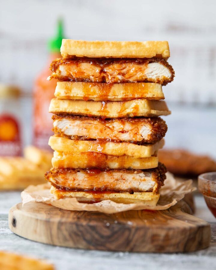 4 cornflake fried chicken waffle sandwiches stacked on each other on wooden board with sriracha maple dripping down