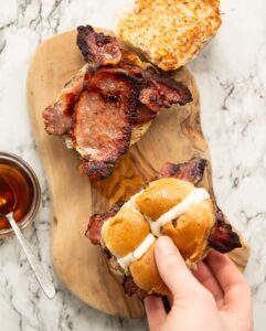 overhead shot of 2 hot cross bun bacon sandwiches with hand holding one on small wooden board