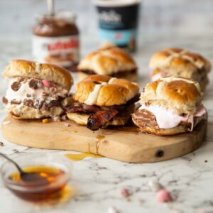 3 hot cross bun sandwiches on wooden board with two more blurred behind surround by garnish
