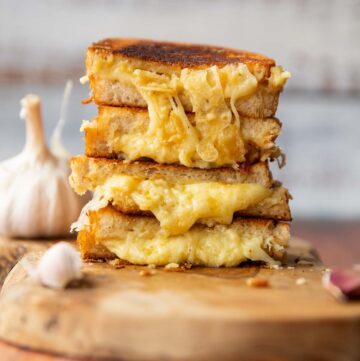 4 roasted garlic grilled cheese halves stacked on each other on wooden board surrounded by garlic