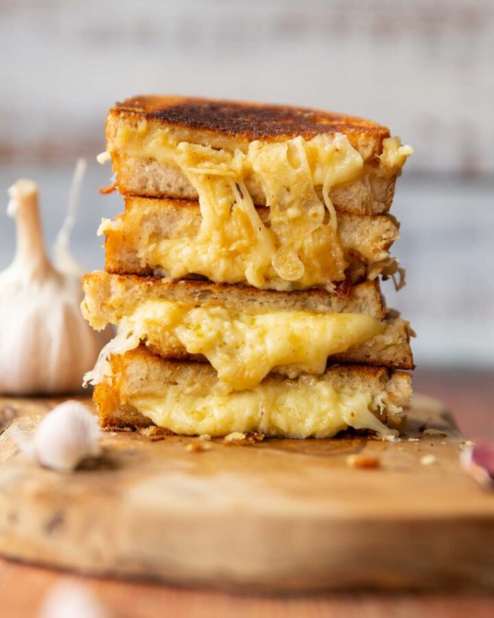 4 roasted garlic grilled cheese halves stacked on each other on wooden board surrounded by garlic