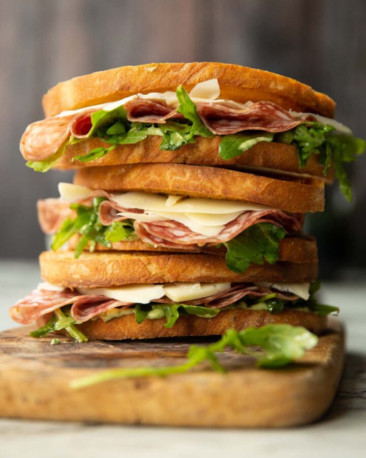 3 salami sandwiches stacked on each other on wooden chopping board