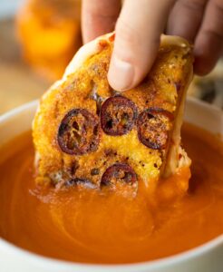 close up shot of hand dunking chilli cheese sandwich into small white bowl of tomato soup
